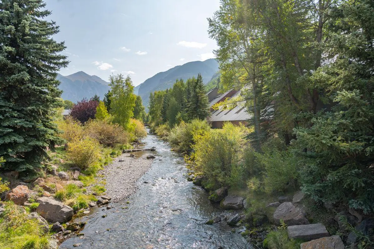 A peaceful scene featuring a flowing creek bordered by lush greenery, with mountains rising majestically in the background. Buildings can be seen nestled beside the water, emphasizing proximity to both nature and amenities. The environment radiates tranquility and offers direct access to hiking, nature walks, and opportunities for photography or fishing.