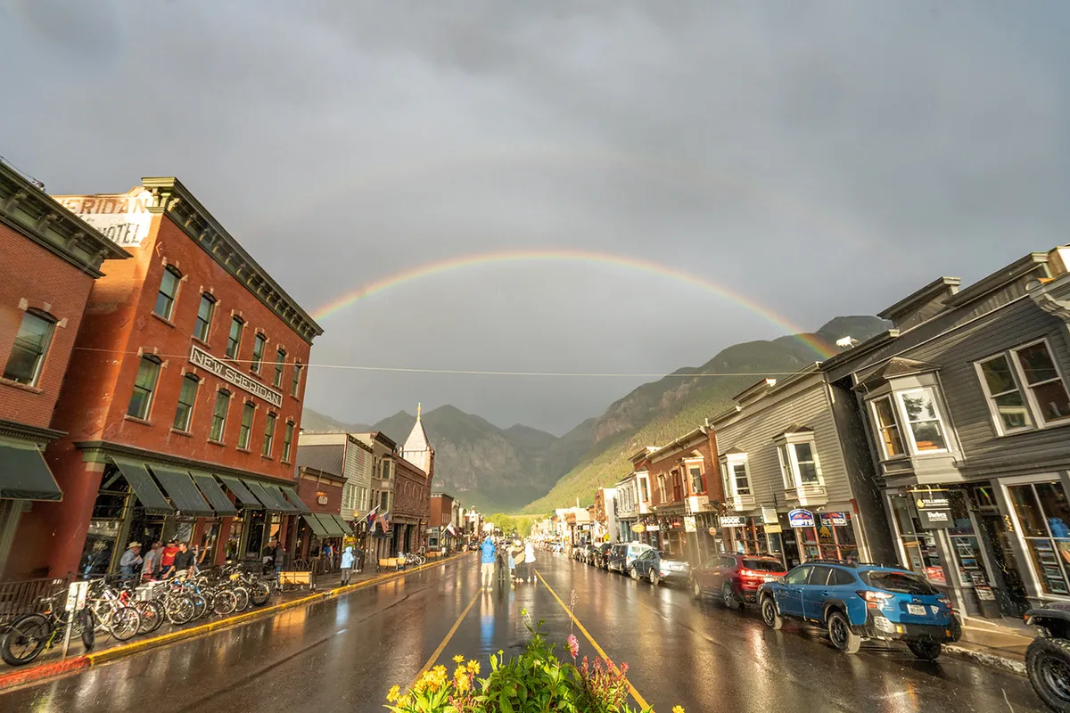 Historic buildings illuminated by a rainbow arching over a wet street, with mountains rising in the background. The scene is lively yet peaceful, suggesting the town's inviting atmosphere after a gentle rain.