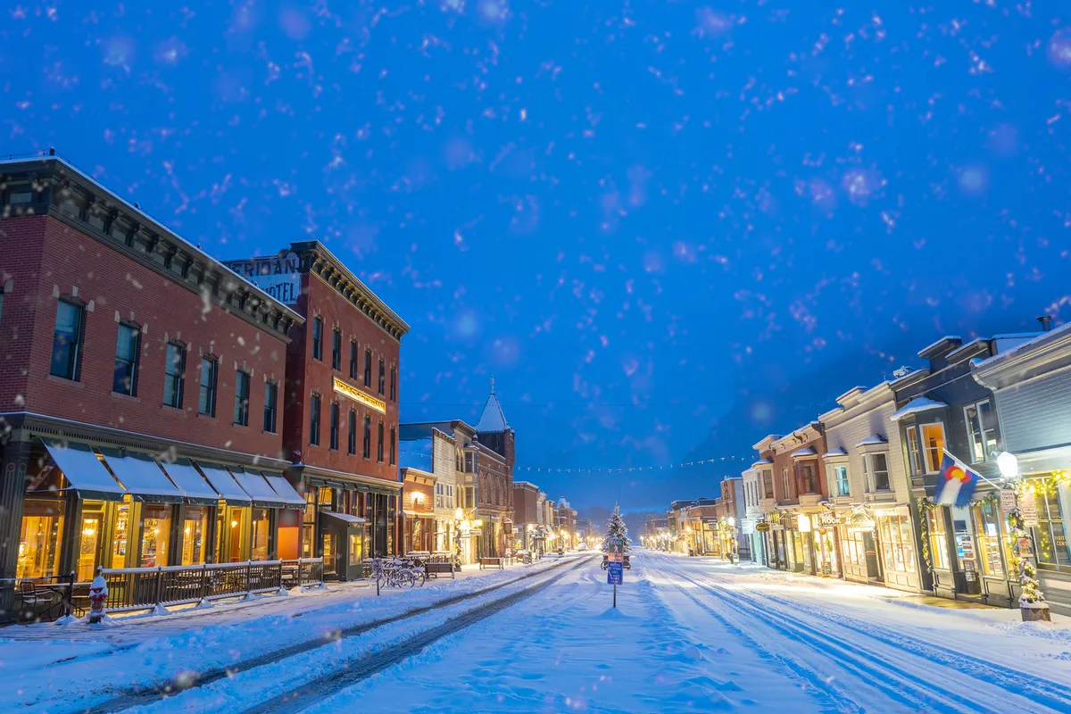 This image depicts a snow-covered street lined with festively decorated buildings, with a Christmas tree twinkling in the distance. The ambiance is warm and inviting, suggesting the charm of Telluride's Main Street in winter. People can be imagined strolling, shopping, or enjoying hot beverages in this cozy, romantic setting.
