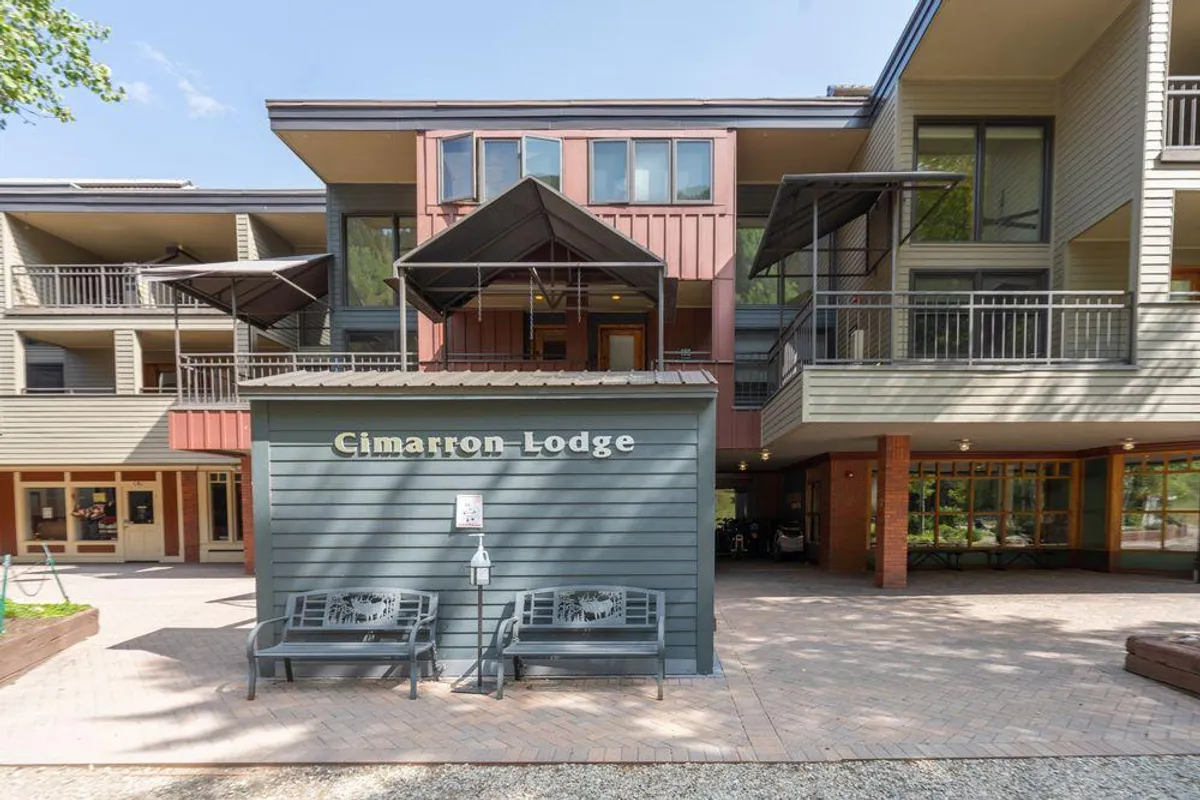 A welcoming exterior view of Cimarron Lodge, showcasing the recognizable lodge sign, benches, and inviting architectural details. The setting suggests a friendly community atmosphere and proximity to ski amenities.