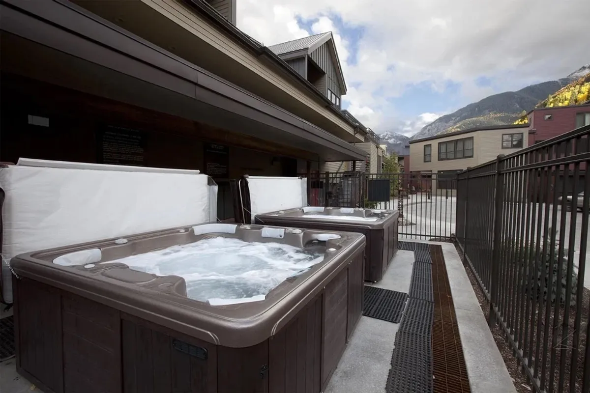 This exterior image features two inviting outdoor hot tubs nestled within a fenced area, surrounded by breathtaking mountain views. The tubs are set against a tranquil backdrop, creating a welcoming space for relaxation and socializing after a day on the slopes.