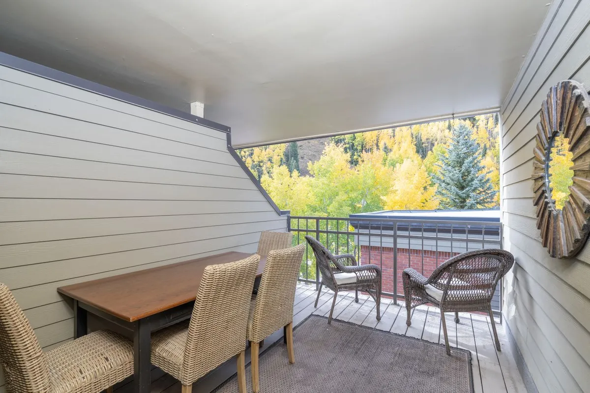 An inviting outdoor dining area features a sturdy wooden table with woven chairs, a wooden railing, and leafy greenery in the background, suggesting a perfect spot for family meals or relaxed conversation.