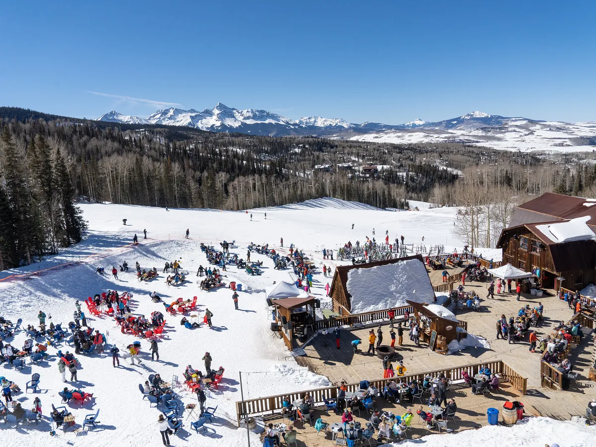 A festive outdoor scene with people skiing and relaxing in a snow-covered seating area, set against a panoramic mountain backdrop. The ambiance is lively, highlighting après-ski socializing and breathtaking scenery.