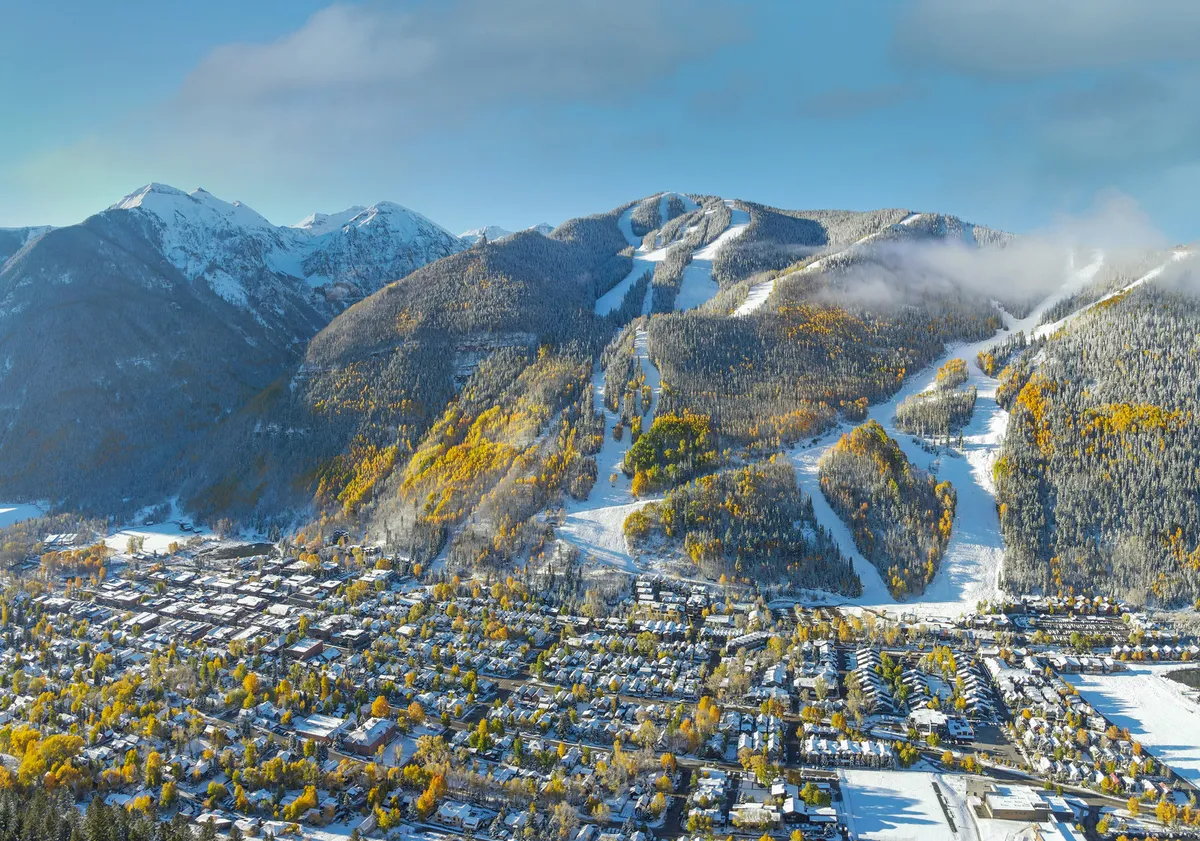A panoramic view of snow-capped mountains, colorful autumn trees, ski slopes, and the town nestled below. The image blends the thrill of mountain adventure with the cozy allure of Telluride’s town amenities.