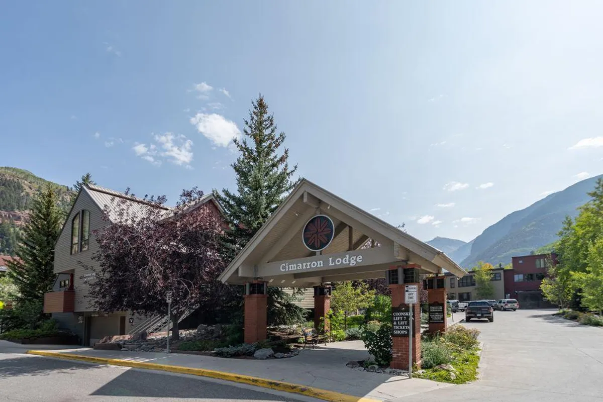 The exterior of Cimarron Lodge is shown with its inviting sign, a landscaped entrance, and mountains in the background. The lodge appears welcoming, highlighting the communal spirit and attractive setting of the property.
