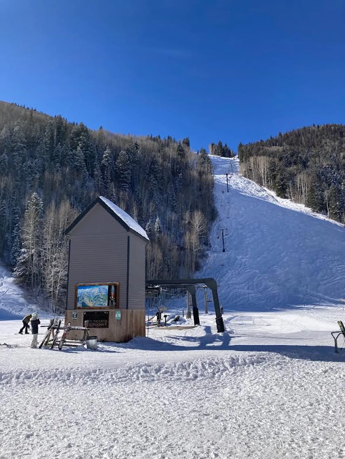 This dynamic exterior scene captures the ski lift and ticket booth at the foot of a snowy slope. Skiers are visible, creating an energetic and cheerful atmosphere that communicates direct access to the mountain and the effortless start to any ski day.