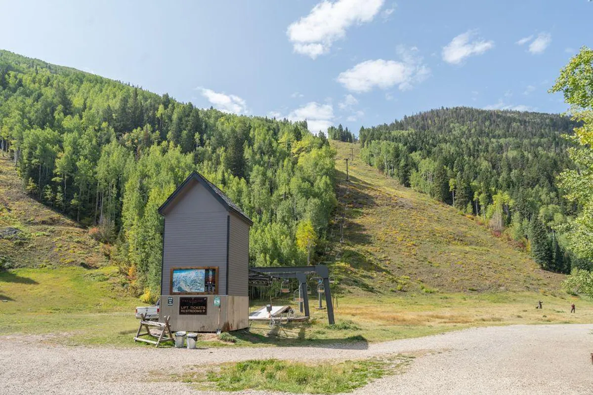 A small building stands beside a ski lift at the base of green, grassy slopes, framed by a mountain backdrop. The area looks peaceful and welcoming, ideal for hiking, picnicking, and gathering with friends during warmer months.