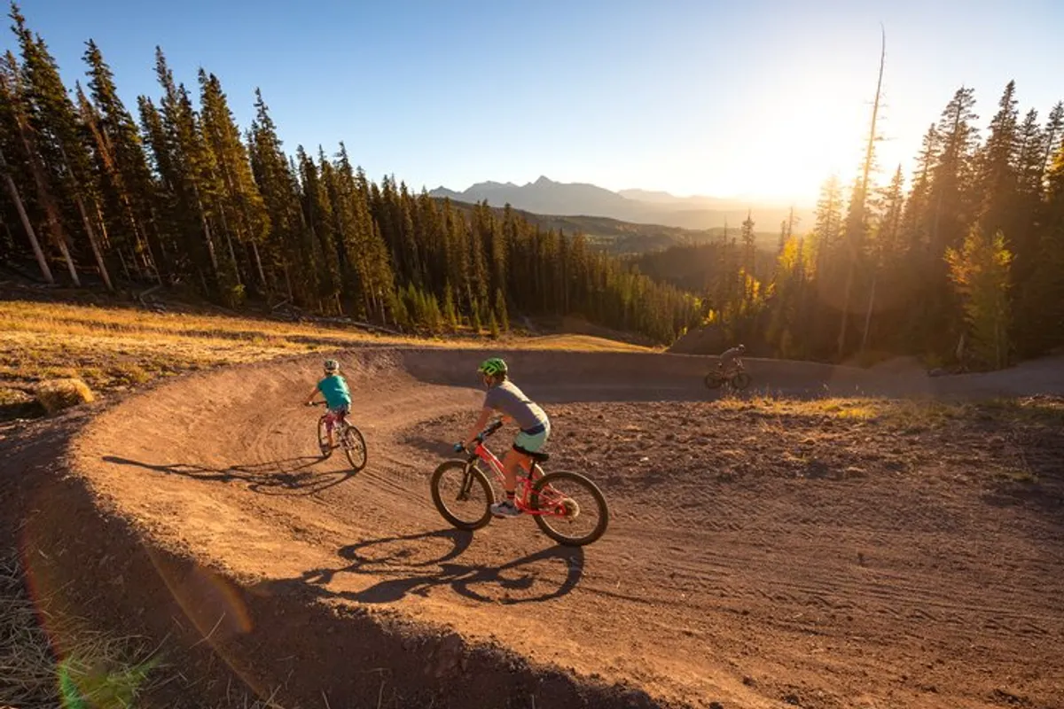 Mountain bike riders navigate a curving dirt path lined with trees, with a dramatic mountain landscape as the backdrop. The scene is filled with energy, emphasizing both the thrill of mountain biking and the camaraderie of enjoying the outdoors together.