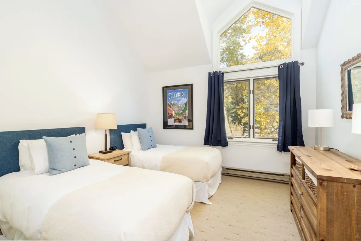 A comfortable guest bedroom with two twin beds, sunlit by a large window. The room also includes a wooden dresser, nightstand with lamp, and framed artwork, creating an inviting environment for families or friends.