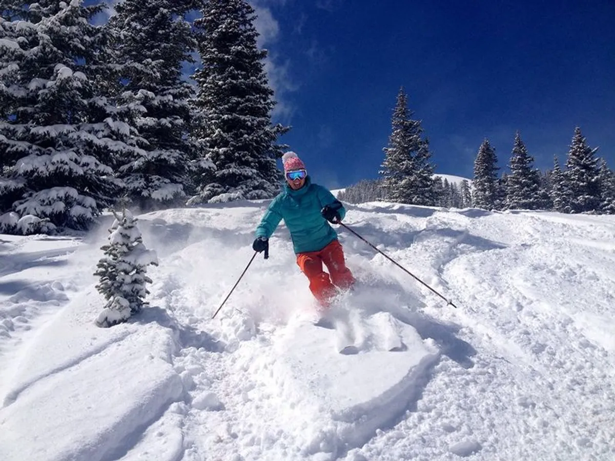 A skier carves down a snow-covered slope, surrounded by tall evergreen trees under a brilliant blue sky. The scene radiates excitement and adventure, with the pristine snow and vibrant winter atmosphere on full display.